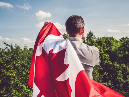 Attractive man holding Canadian Flag on blue sky background on a clear, sunny day. View from the...