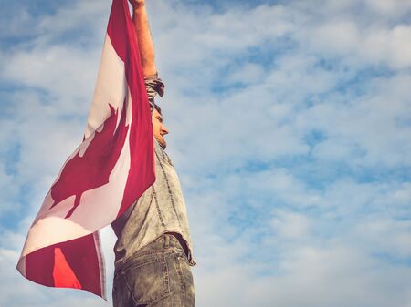 Attractive man holding Canadian Flag on blue sky background on a clear, sunny day. View from the back, close-up. National holiday conceptの写真素材