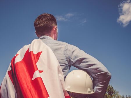 Young engineer, white hardhat and Canadian Flag in the park against the backdrop of green trees and...