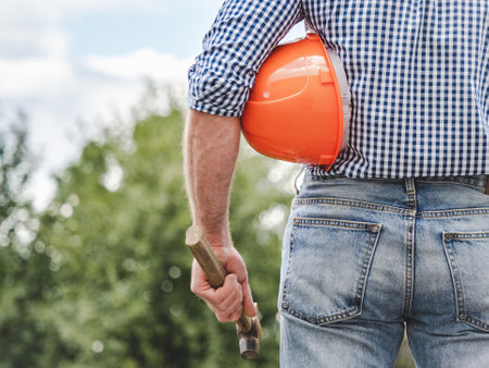 Man in work clothes with a protective helmet against a background of trees and blue sky. Rear view,...