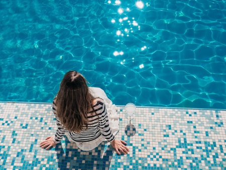 Stylish woman with a glass of drink sits near the swimming pool. Sunny morning, clear day. Top view....
