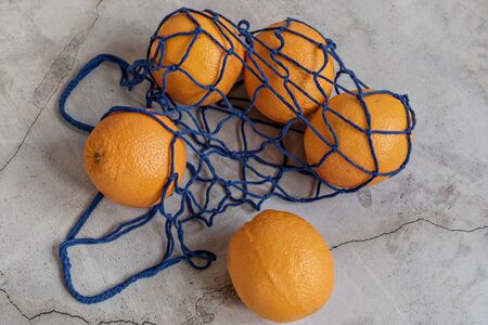 Oranges in an eco-netted blue bag on a gray background. Environmental care, waste recycling