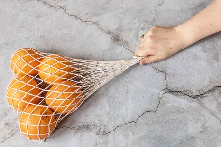 A woman's hand holds a white string bag with oranges on a dark background. Environmental care, waste recyclingの写真素材