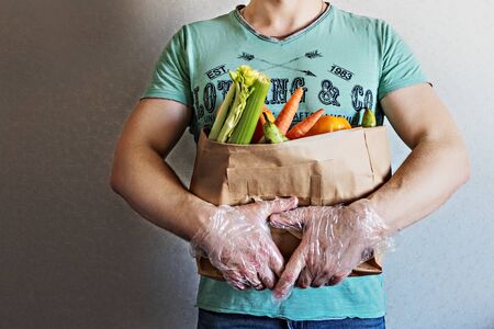 A man holds an eco paper bag with groceries in his hands. Food delivery to the door to the house by courier. Hands in gloves. Coronavirus, COVID 19の写真素材