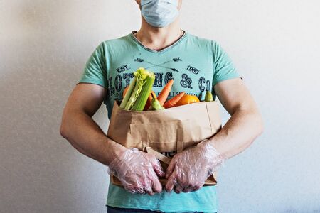 A man holds an eco paper bag with groceries in his hands. Food delivery to the door to the house by courier. Hands in gloves. Coronavirus, COVID 19の写真素材