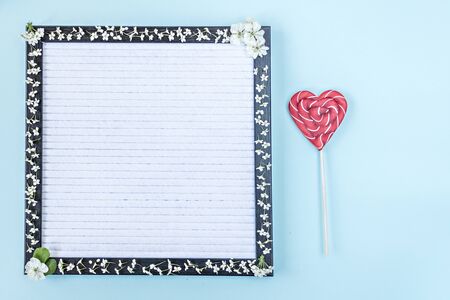 Candy on a stick in the shape of a heart on a writing board, which is decorated with white small flowers. Place for textの写真素材