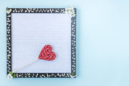 Candy on a stick in the shape of a heart on a writing board, which is decorated with white small flowers. Place for textの写真素材