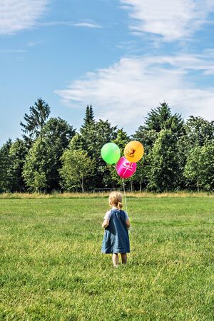 A little girl with colorful balls standing with his back and looks towards the forest. On the balloon inscription happy birthdayの写真素材