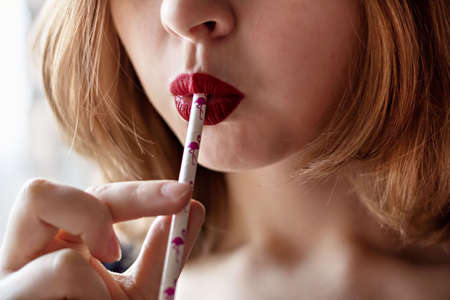 A young girl with bright red lipstick drinks a cocktail from a glass from a paper tube. The concept of ecology and environmental protectionの写真素材
