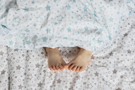 Close-up of toddler girl's feet on the bed under the blanket. Light blue and beige tonesの写真素材