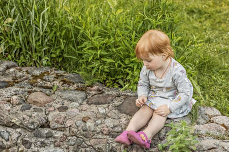 Little girl with red hair sits with a camomile in her hand in the garden among the greenery. Selective focus.の写真素材