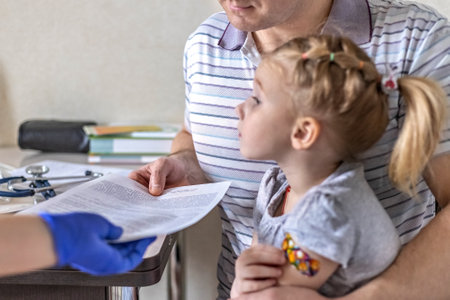 Little girl with her father in the doctor's office at the clinic. Vaccinated against coronavirus. The doctor passes the documents to the girl's dad. The concept of vaccination from Covid-19.のeditorial素材