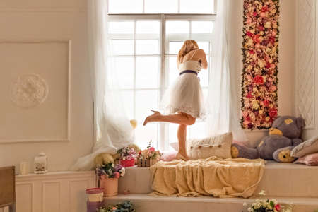 Portrait of a young woman in a white dress against the background of a closed large window, looking out into the street through the glass.の写真素材