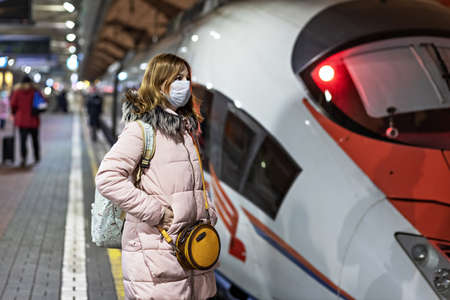 A young woman in a medical mask stands on the platform of the station waiting for the departure of a...