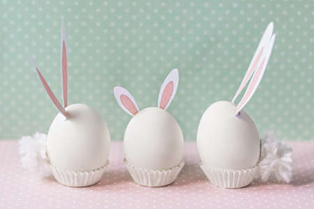 White chicken eggs with bunny ears and tails on bed flowers background. A family. Happy Easter holiday concept.の写真素材