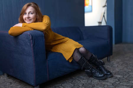 Portrait of a smiling woman with red hair resting her head on her hands on the back of the sofa.の写真素材