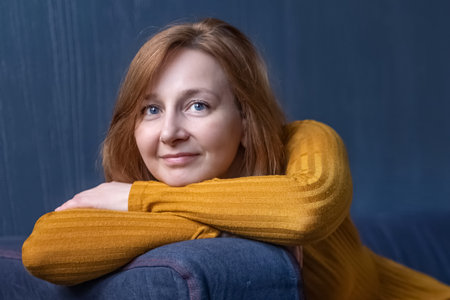 Portrait of a smiling woman with red hair resting her head on her hands on the back of the sofa.