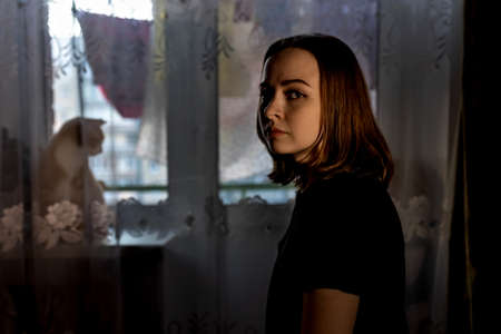 Portrait of a young teenage girl in her room in the evening. Red cat at the window, silit on the windowsill.の写真素材