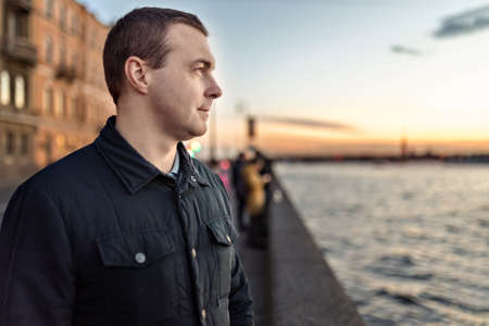 Portrait of a young man watching the sunset on the embankment of the river in the city.の写真素材