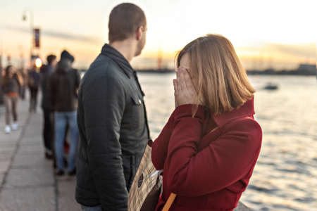Young couple man and woman in a quarrel, on the embankment of the river in the city at sunset. The girl covered her face with her hands, crying. Emotions.の写真素材