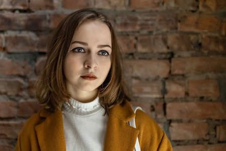 Portrait of a young woman in a bright yellow coat with a serious face against a brick wall background.の写真素材