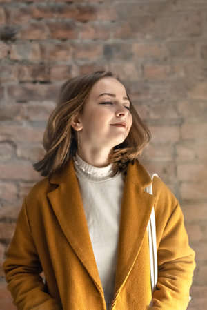 Portrait of a young woman in a bright yellow coat against the background of a brick wall by the window.In the sunshineの写真素材