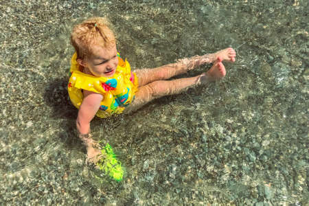 A little girl toddler is sitting in a vest and a water pistol in the water on the seashore.の写真素材