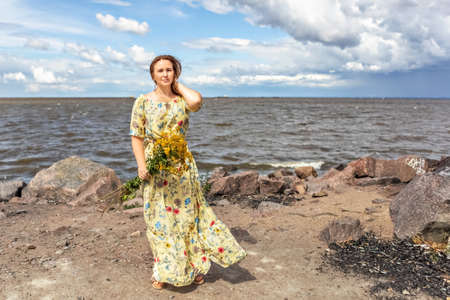A young woman in a yellow dress with a bouquet of tansy stands on the beach in the wind. Light and airy.の写真素材