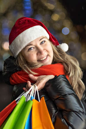 Portrait of a woman in a Santa hat with Christmas shopping in colorful bags on the street decorated with Christmas lights and decorations.の写真素材