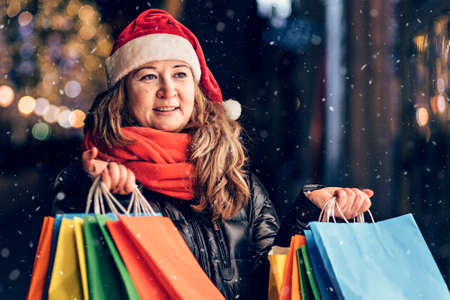 Portrait of a woman in a Santa hat with Christmas shopping in colorful bags on the street decorated with Christmas lights and decorations.の写真素材