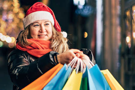 Portrait of a woman in a Santa hat with Christmas shopping in colorful bags on the street decorated with Christmas lights and decorations.の写真素材
