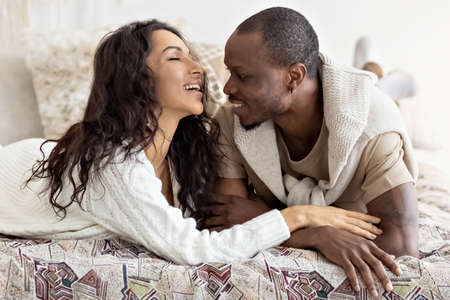Happy African American man and caucasian woman in warm sweaters lying on bed at home, looking at...