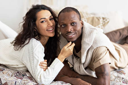 Happy interracial couple looking at camera while lying on bed at homeの写真素材