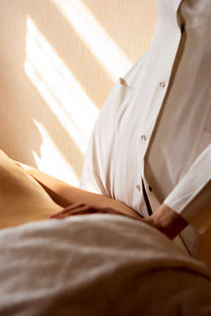 Top view of a professional female masseur with strong arms massaging the back and shoulders of a muscular athletic man lying on his stomach at a massage table in a luxury salon, against the backdrop of sunlight.の写真素材