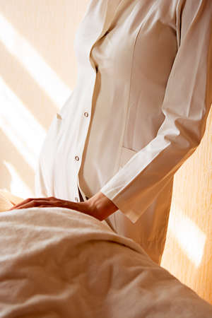 Top view of a professional female masseur with strong arms massaging the back and shoulders of a muscular athletic man lying on his stomach at a massage table in a luxury salon, against the backdrop of sunlight.の写真素材