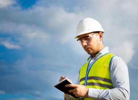 engineer in a helmet, glasses and a yellow vest, hand on a helmet, holding a notebook, a folder, making notes in a notebook a caucasian white engineerの写真素材