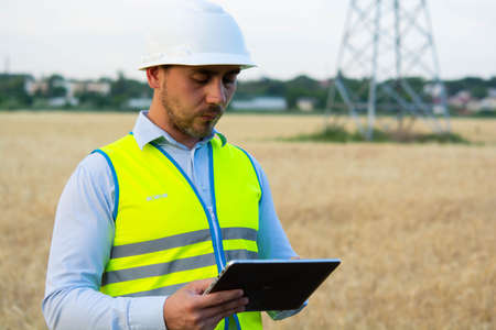engineer in a helmet, glasses and a yellow vest, hand on a helmet, holding a notebook, a folder,...
