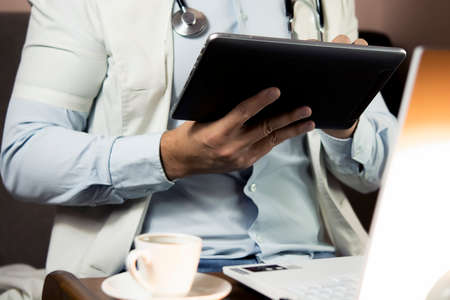 Serious professional doctor in white coat and stethoscope holding modern touch screen gadget using digital tablet computer at work. The concept of a medical network of data transmission and telemedicine in the field of healthcare.の写真素材