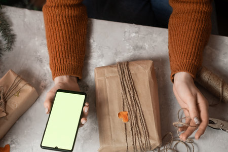 a young woman holds a phone, a tablet in her hand and fulfills an order by phone, taking an order for gift wrapping, in the background a Christmas tree and lights, the concept of preparing for the new year, merry christmas, new year 2023の写真素材