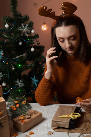 a happy young woman makes an order by phone, the girl has New Year's deer antlers on her head, taking an order for gift wrapping, in the background a Christmas tree and lights, the concept of preparing for the new year, merry christmas, new year 2023の写真素材