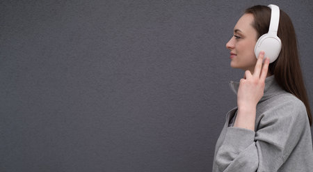 Carefree attractive brunette girl, dressed in a gray sweater, listening to music on headphones, standing on a gray backgroundの写真素材