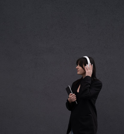 A beautiful girl in white headphones and a black jacket holds a tablet outside, work outside, listen to music and sing along, office workerの写真素材