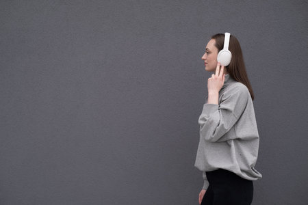 Carefree attractive brunette girl, dressed in a gray sweater, listening to music on headphones, standing on a gray backgroundの写真素材