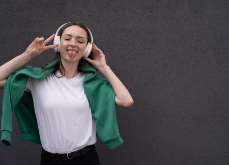 Emotional girl listens to music in headphones on a gray background, shows tongue and heavy metal sign with hands. Expressive girl listening to rock music with headphonesの写真素材