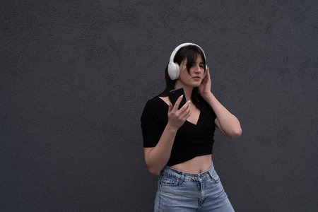 Smiling caucasian young woman listening to rock band singer music song e-book podcast in headphones choosing audio track on mobile phone on gray backgroundの写真素材