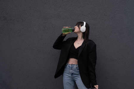 Young beautiful smiling woman, with headphones around her neck and a bottle in her hand, looks at the camera, on a gray backgroundの写真素材