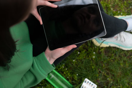 Mockup image of woman sitting and holding black tablet with blank white screen on green grass in parkの写真素材