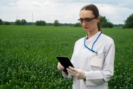 a female agronomist in a white coat checks the growth of plants in the field. A biologist in the field checks the readings of wheat, a farmer takes notes on a tablet, agribusiness conceptの写真素材