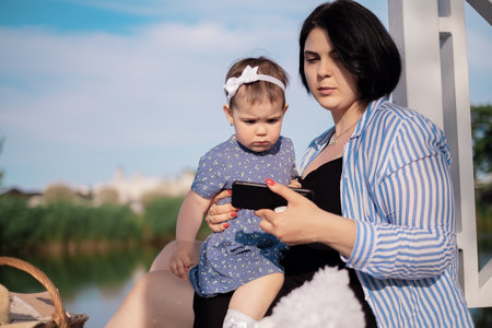 Mom and daughter on a picnic by the lake eat and watch cartoons on the phoneの写真素材