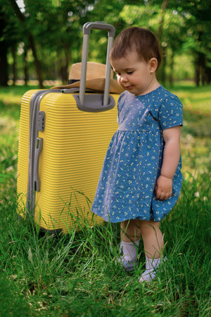 A little happy girl in a blue summer dress stands next to a suitcase and green plants and laughs. Summer vacation, tourism, vacation trip, summer vacation, sea tripの写真素材